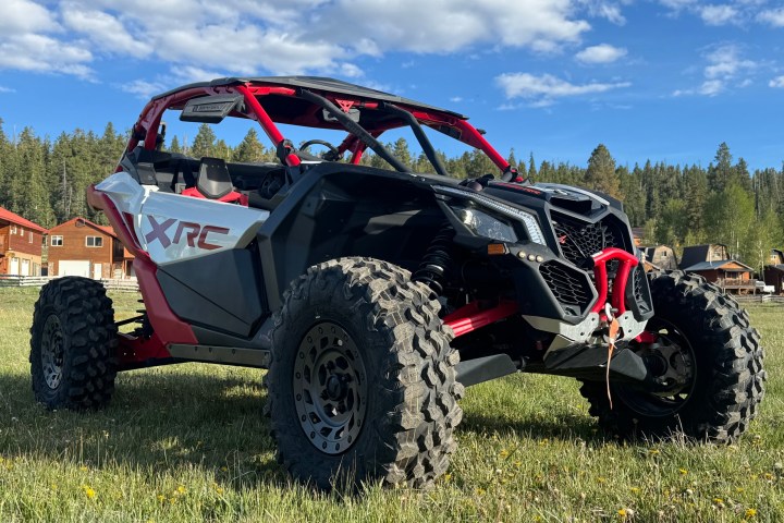 Red and black off-road vehicle on grassy field with trees and buildings in background.